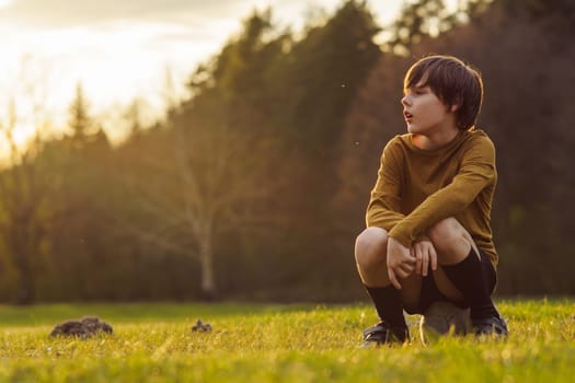 In early summer morning, teenager in long-sleeved T-shirt sits on soccer ball on field waiting for workout. Sports and health. Children's sports sections. Hobbies and hobbies. Summer sports camp.