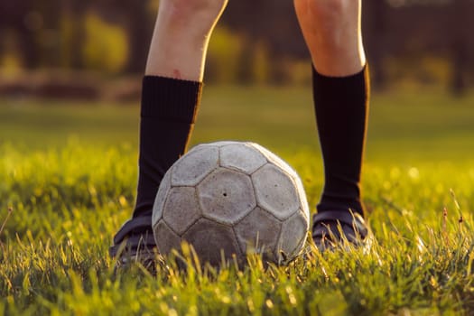 Unrecognizable young athlete in high socks stands in front of soccer ball on mown grass in early summer morning. Sports and health. Children's sections. Football practice. Summer sports camp.