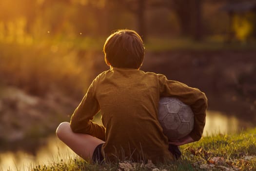 Teenager in long-sleeved T-shirt and ball under his arm sits with his back to camera after training on field and looks at stream. Children's sports sections. Football practice. Summer sports camp.