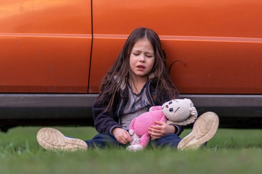 Children's problems. Little lonely girl is very upset, tired, holding toy in her hands, sitting on grass with her back pressed against car. Child is upset and offended. Inner world of child.