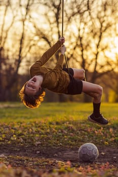 Happy childhood. Smiling teenage boy is hanging upside down on homemade swing in nature, in park, in village, in children's camp in rays of sunset. Outdoor games and entertainment. Child protection.