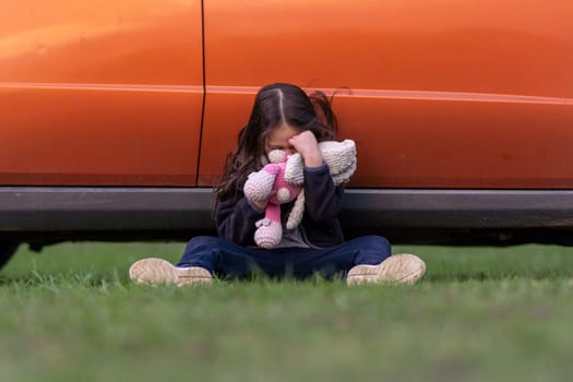 Little lonely girl is crying, hiding her face in toy, sitting on grass with her back pressed against car. Children's tears. Child is upset and offended. Children's problems. Inner world of child.