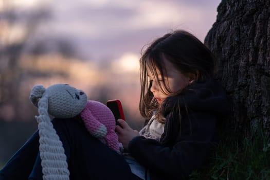 Charming little girl with toy on her lap is leaning against tree, enthusiastically looking at her phone at sunset in village. Happy childhood. Summer holidays. Summer in country. Time to dream.