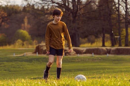 Teenage boy of 11-13 years old in sports uniform trains early in summer morning on field with soccer ball against background of stacks of mown grass. Sports and health. Sports activities .
