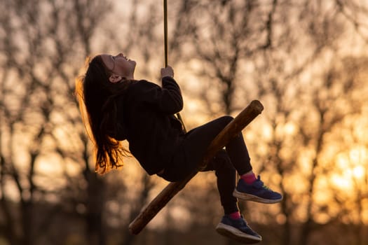 Portrait of happy little girl who swings on homemade swing in nature, in park, in village, in children's camp against background of trees. Outdoor games and entertainment. Child protection.