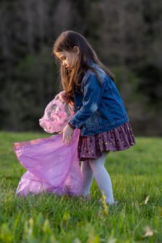 Vertical portrait of charming little girl with long hair in denim jacket collecting garbage in nature, in meadow on summer day. Environmental problems. We take care of nature. Saving planet.