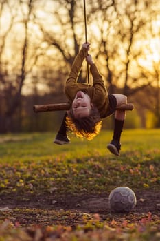 Child protection. Smiling teenage boy is hanging upside down on homemade swing in nature, in park, in village, in children's camp in rays of sunset. Outdoor games and entertainment. Happy childhood.