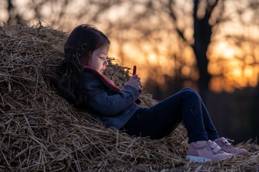Beautiful portrait of charming little girl lying on haystack playing with her phone against background of setting sun. Happy childhood. Summer in country. Time to dream. Inner world of child.