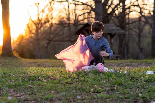 Children's volunteer activity. Teenage boy collects garbage in large pink bag in nature, in meadow on autumn evening at sunset. Environmental problems. We take care of nature. Saving planet.