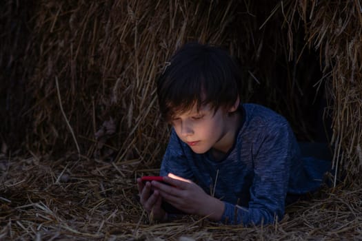 Boy lies in haystack on summer night and reads something exciting from phone screen. Happy childhood. Summer holidays. Summer in country. Time to dream. Inner world of child. Kids are outside.