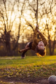 Smiling teenage boy is hanging upside down on homemade swing in nature, in park, in village in rays of sunset. Outdoor games and entertainment. Happy childhood. Summer in country. Child protection.