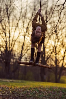 Happy teenage boy hanging upside down on homemade swing in nature, in park, in village in sunset rays in evening. Positive emotions. Child protection. Child safety. Autumn portrait.