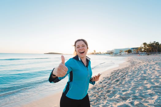 Smiling female runner show thumbs up. Summer day on the beach.