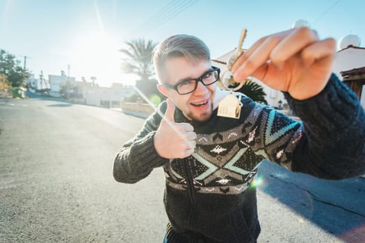 Happy funny man holds house keys on house shaped keychain in front of a new home.