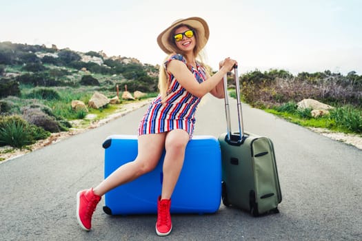 Happy traveler woman sits on vintage suitcase on road.