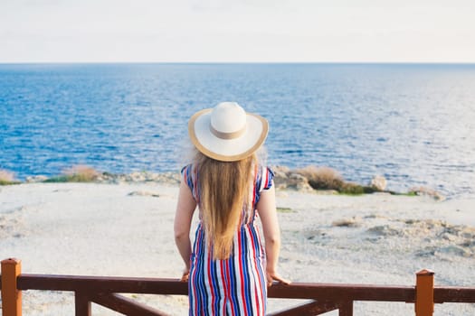 Closeup back view of woman sitting in white dress and hat looking out towards blue ocean and sky, isolated sea background