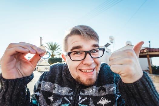 Happy funny man holds house keys on house shaped keychain in front of a new home.