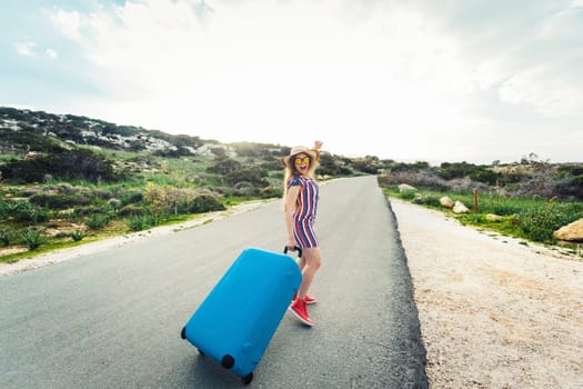 Traveler woman wearing a dress and hat sits on blue suitcase on the road and laughs .
