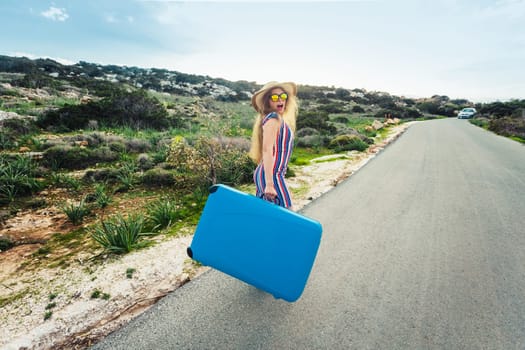 Traveler woman wearing a dress and hat sits on blue suitcase on the road and laughs .