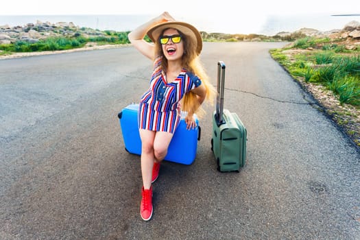 Traveler woman wearing a dress and hat sits on blue suitcase on the road and laughs .