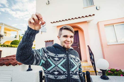 Happy funny man holds house keys on house shaped keychain in front of a new home.