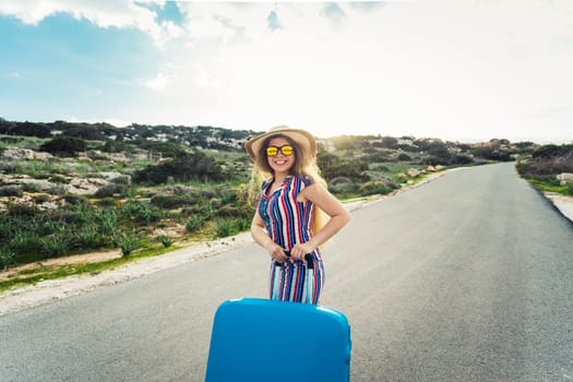 Happy smiling woman with blue suitcase on a road.