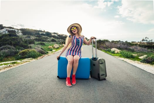 Happy traveler woman sitting on a suitcase on the road and smiling. Concept of travel, journey, trip.