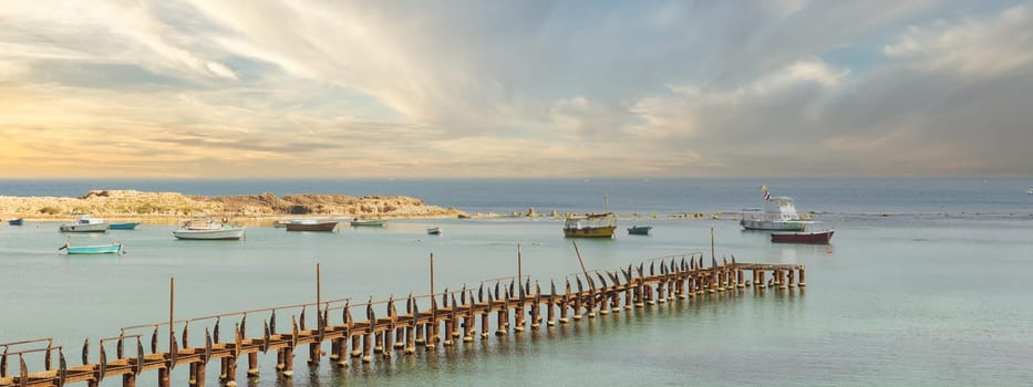 Abandoned ruined fishing pier by the sea in Alexandria, Egypt, with colorful fishing boats in the far end. The pier is framed by the azure waters of the Mediterranean Sea after sunrise
