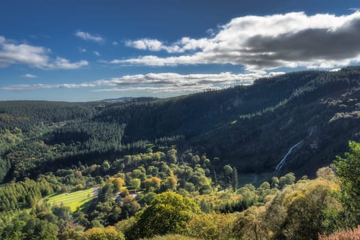 Tall Powerscourt Waterfall, valley, forests in autumn colours illuminated by sunlight. Scenic Wicklow Mountains, Ireland