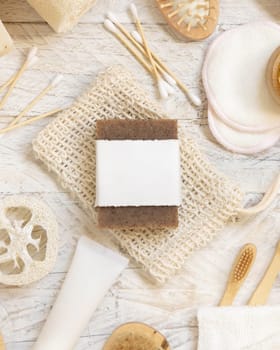 Organic handmade beige soap bar with blank label on soap saver bag near hygiene Items - natural sponges and brush, bamboo toothbrushes and cotton swabs, top view, mockup