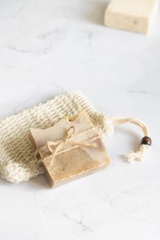 Beige handmade soap bar on sisal soap saver bag on white marble table close up, copy space. Natural herbal products for face and body care. Organic handmade skincare products