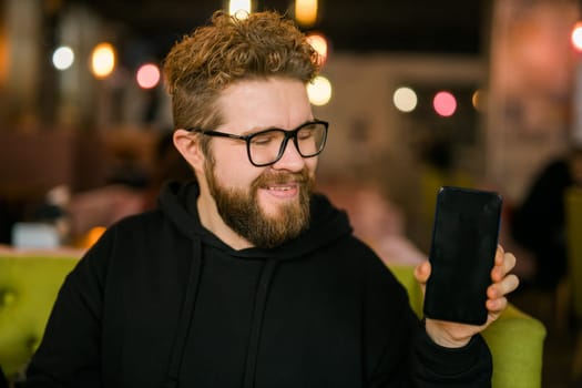 Satisfied man with dark beard holding smartphone and showing his favorite mobile app Male programmer has developed a cool application