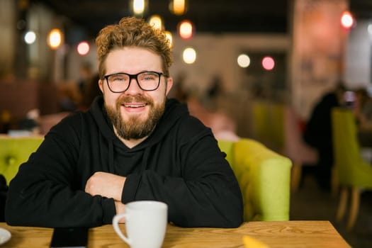 Bearded curly man smiling confident drinking coffee in restaurant or coffee shop. Millennial generation and Gen Y.