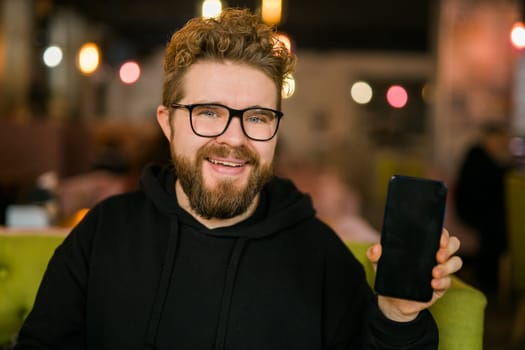 Satisfied man with dark beard holding smartphone and showing his favorite mobile app Male programmer has developed a cool application