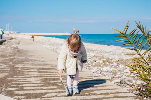 Child, childhood and people concept - Baby girl sits on asphalt square road and play with stick near sea beach