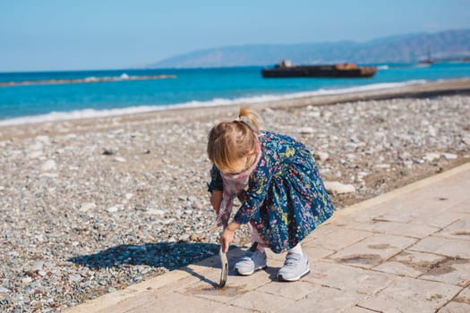Child, childhood and people concept - Baby girl sits on asphalt square road and play with stick near sea beach