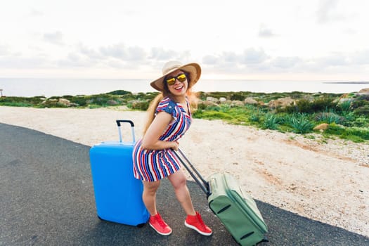 Happy traveler woman sitting on a suitcase on the road and smiling. Concept of travel, journey, trip.