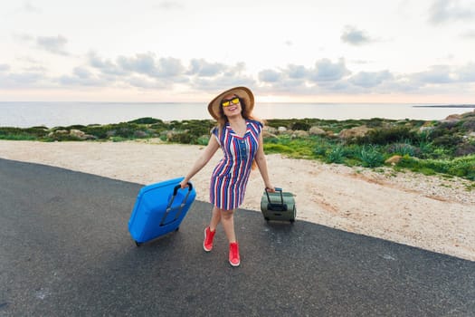 Happy traveler woman sitting on a suitcase on the road and smiling. Concept of travel, journey, trip.