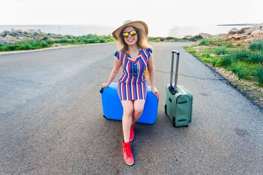 Happy traveler woman sitting on a suitcase on the road and smiling. Concept of travel, journey, trip.