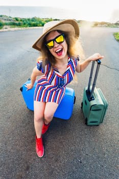 Happy traveler woman sitting on a suitcase on the road and smiling. Concept of travel, journey, trip.
