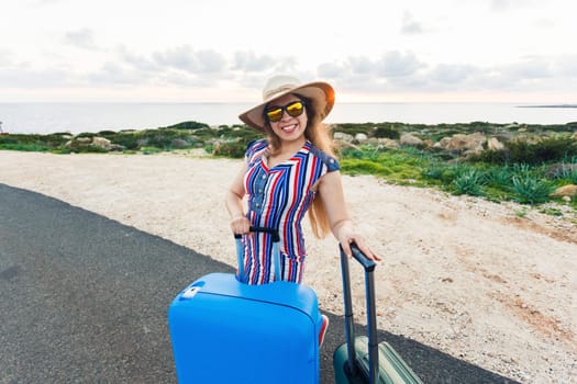 Happy traveler woman sitting on a suitcase on the road and smiling. Concept of travel, journey, trip.