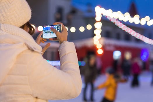 A woman on the rink is skating and taking selfie on smartphone. New Years Eve and Christmas. Fairy lights. Ice and snow mood concept. Winter