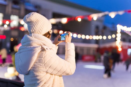 A woman on the rink is skating and taking selfie on smartphone. New Years Eve and Christmas. Fairy lights. Ice and snow mood concept. Winter