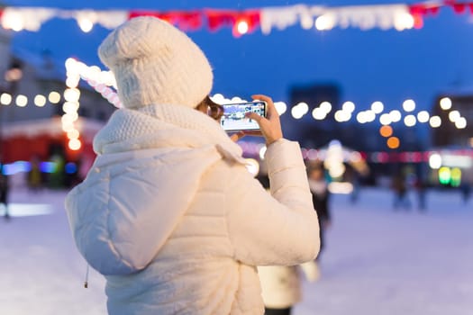 Christmas, winter, technology and leisure concept - happy young woman taking picture with smartphone on ice skating rink.
