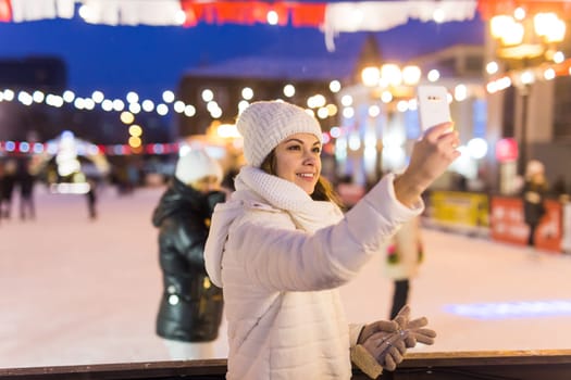 A woman on the rink is skating and taking selfie on smartphone. New Years Eve and Christmas. Fairy lights. Ice and snow mood concept. Winter