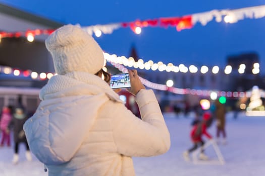 Christmas, winter, technology and leisure concept - happy young woman taking picture with smartphone on ice skating rink.