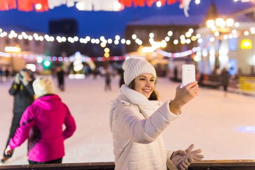 A woman on the rink is skating and taking selfie on smartphone. New Years Eve and Christmas. Fairy lights. Ice and snow mood concept. Winter
