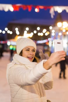 A woman on the rink is skating and taking selfie on smartphone. New Years Eve and Christmas. Fairy lights. Ice and snow mood concept. Winter