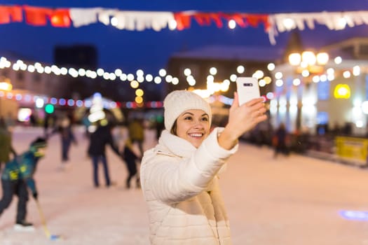 Happy young woman in winter on the ice rink taking picture on smartphone.