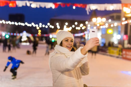 Christmas, winter, technology and leisure concept - happy young woman taking picture with smartphone on ice skating rink.